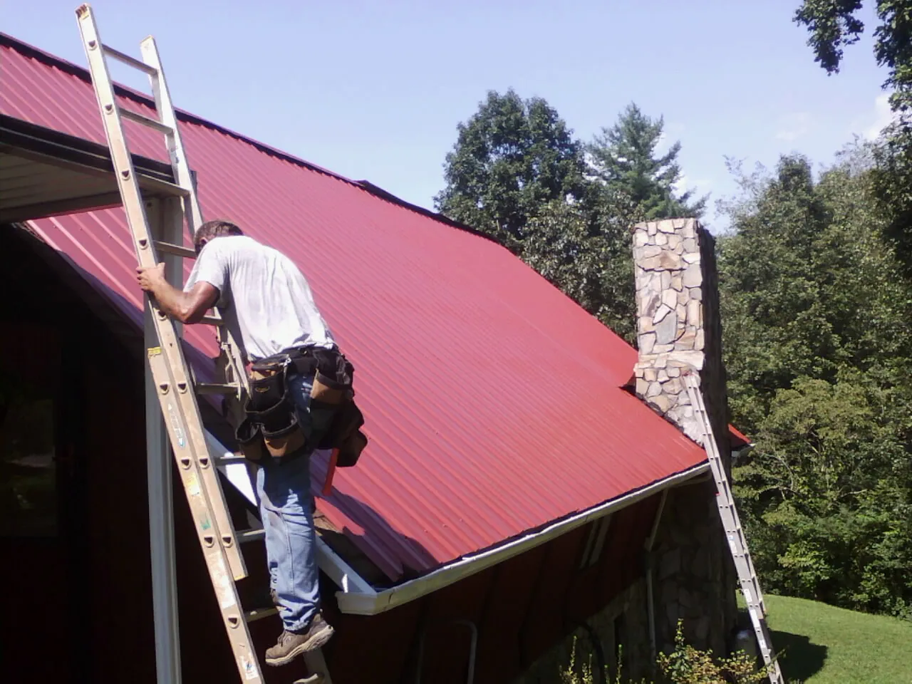 Roofer on a ladder beside a red metal roof and stone chimney — Soffit & Fascia Repair by Roofing Brothers in Thomaston